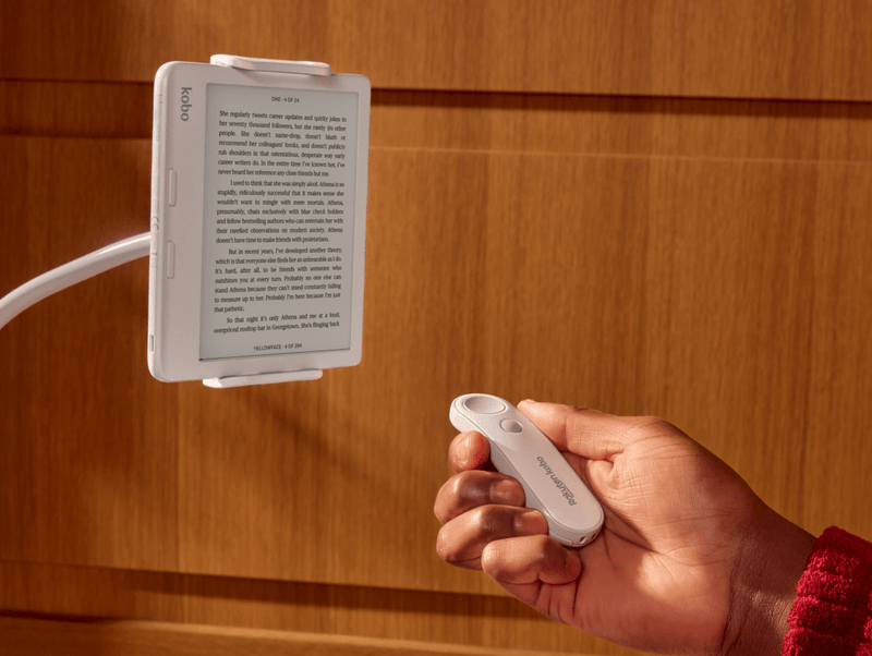 A white Kobo Remote is shown in someone's right hand, pointing towards a white Kobo Libra Colour eReader on a bendable stand.