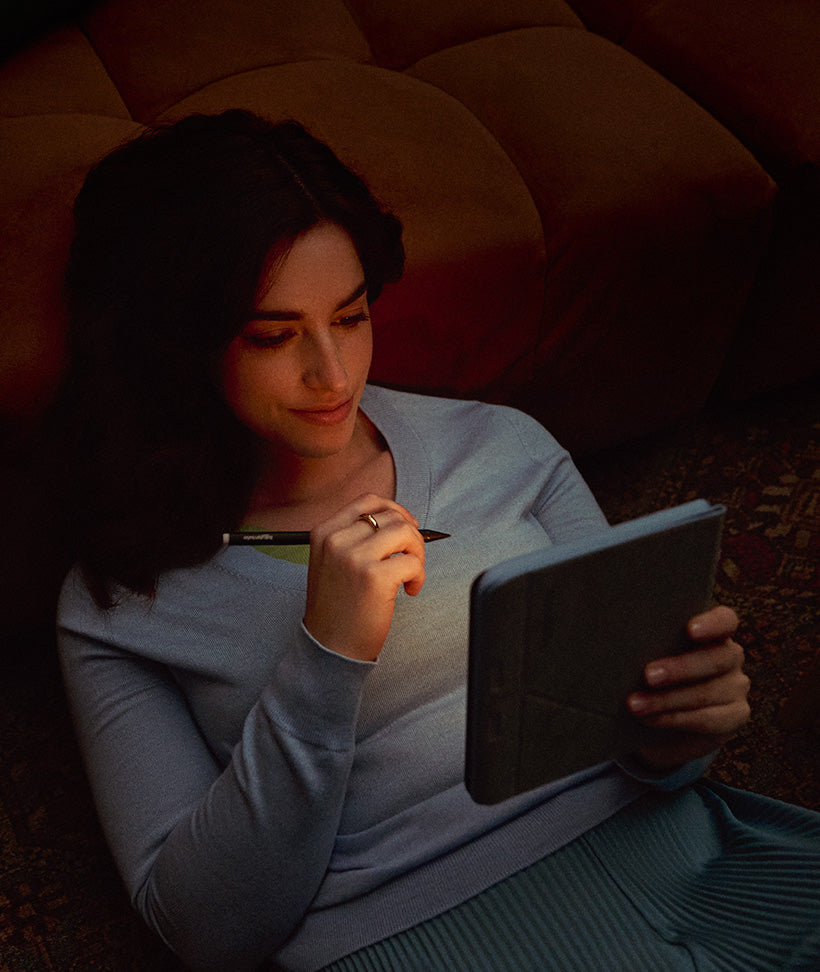 A woman reclining on a couch, lit by the soft light of a Kobo Libra Colour eReader she is holding, along with a Kobo Stylus 2.